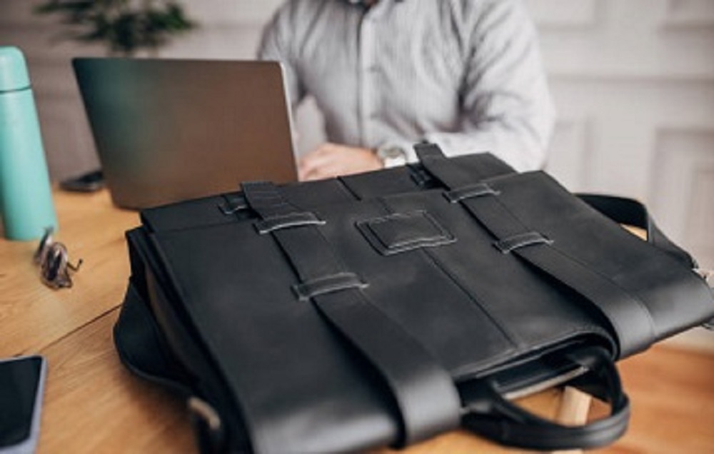 Unrecognizable elegant businessman sitting at desk and using laptop, in front of him is modern leather bag.